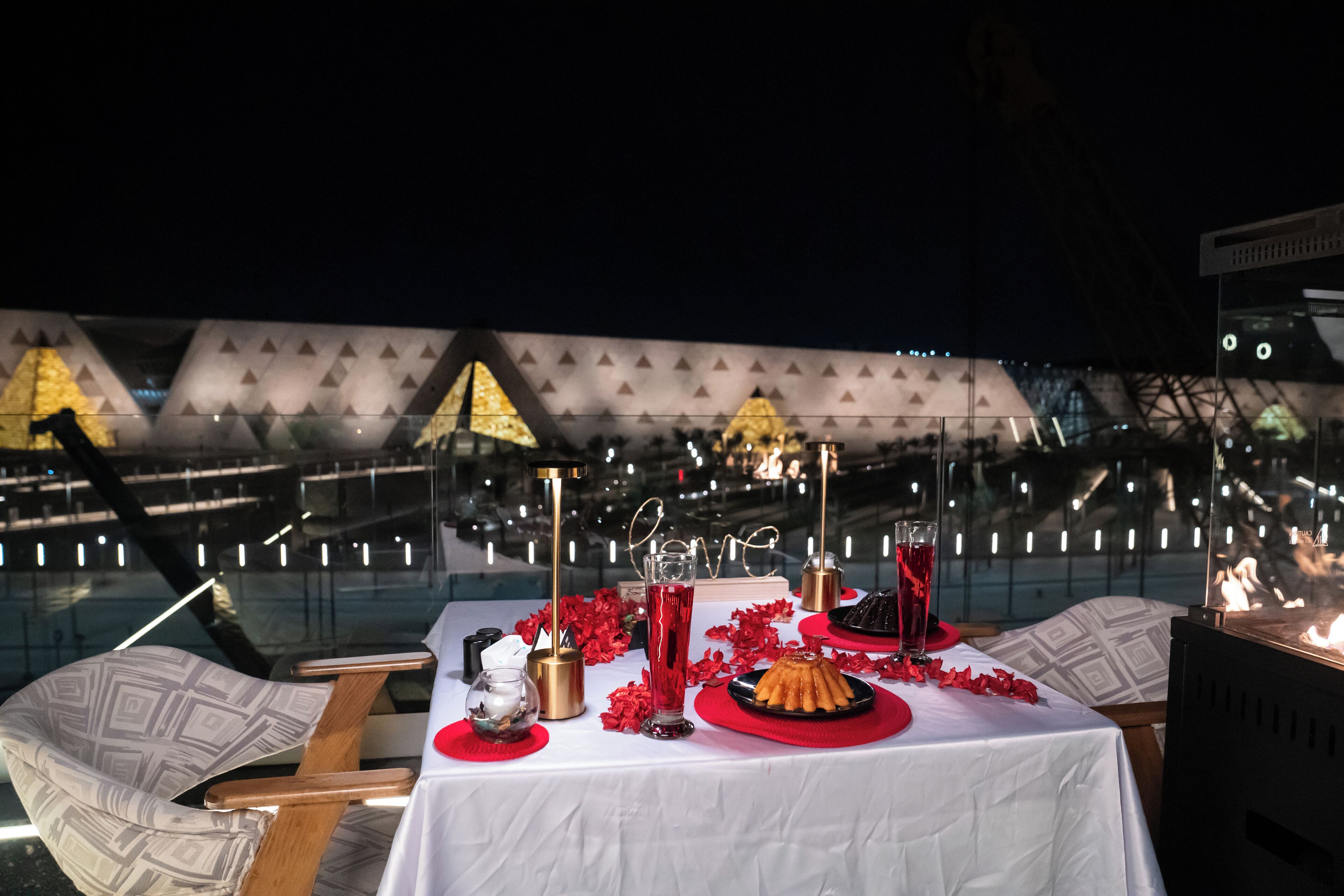 Une table dressée avec une nappe blanche, des serviettes rouges et une assiette à dessert, avec en arrière-plan la vue d’un bâtiment aux motifs triangulaires.