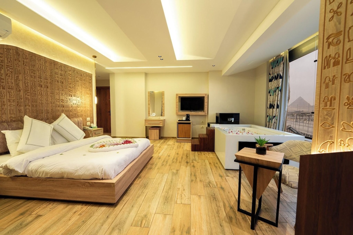 A modern hotel room with a wooden floor, a large bed with white bedding, a desk with a computer, a TV mounted on the wall, and a large window with a view of a mountain.