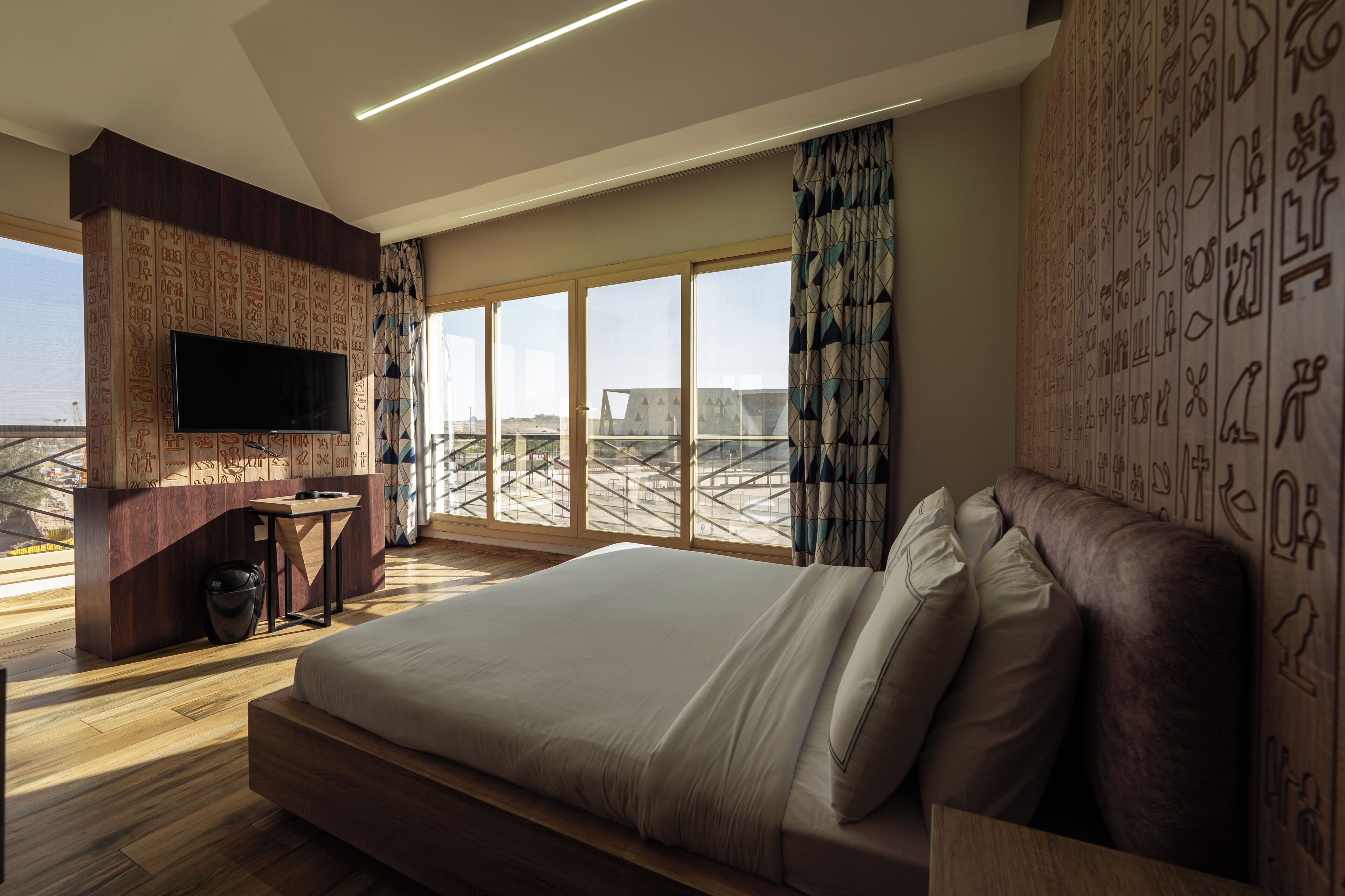 A modern hotel room with a large bed, a flat-screen TV mounted on a wall with hieroglyphs, a small table, and a view of the cityscape through large windows.