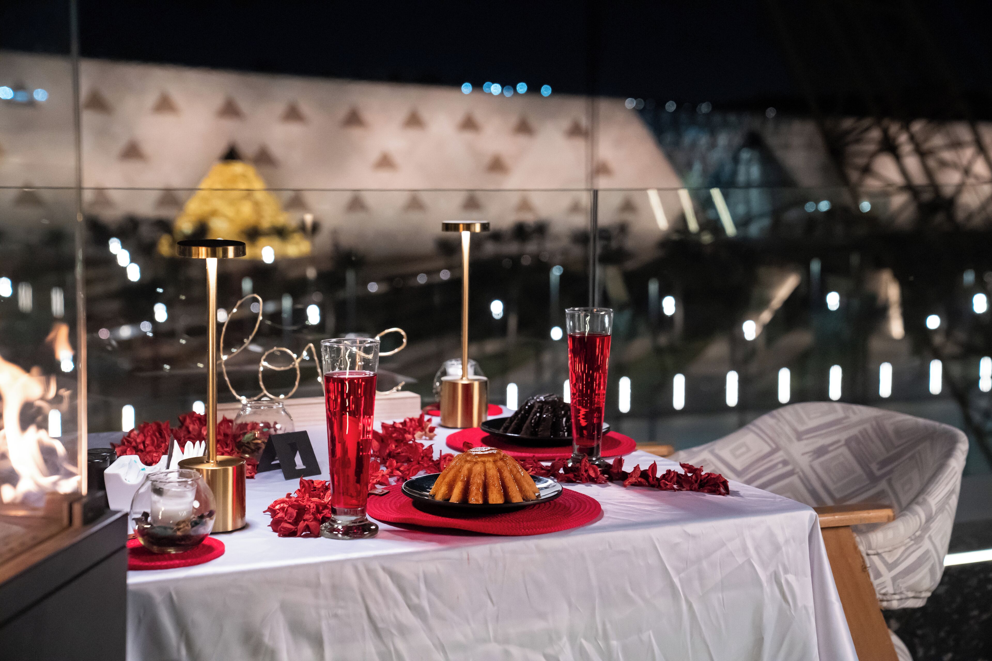 A table set for a meal with a dessert, glasses, and a candle holder, with a view of a building in the background.