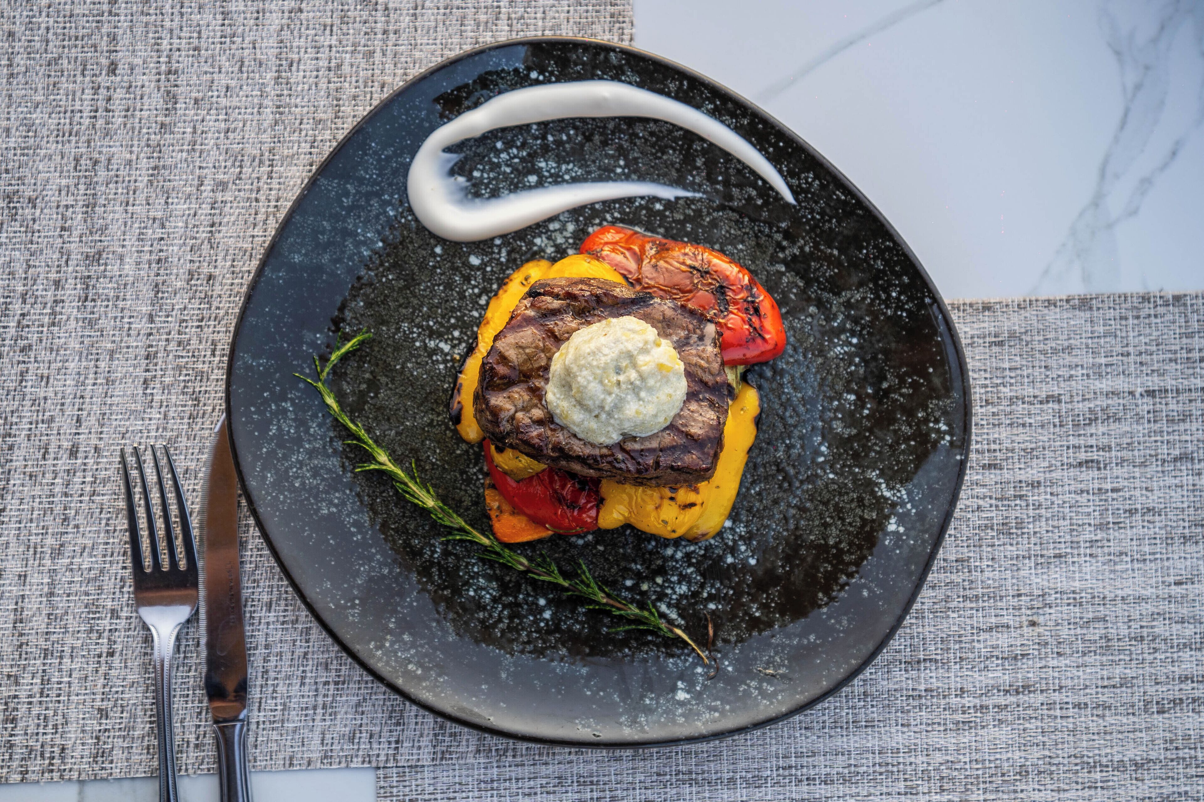 Un plato con carne a la parrilla, verduras y una cucharada de salsa sobre un plato oscuro.