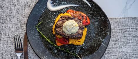 Un plato con carne a la parrilla, verduras y una cucharada de salsa sobre un plato oscuro.