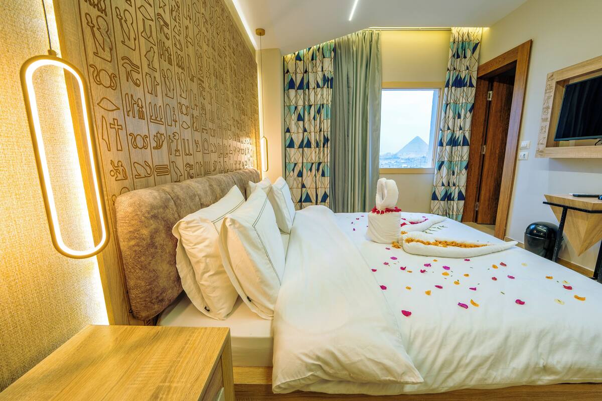 A hotel room with a bed, pillows, a bedside table, a wall with hieroglyphs, a window with curtains, and a TV on a wooden stand.