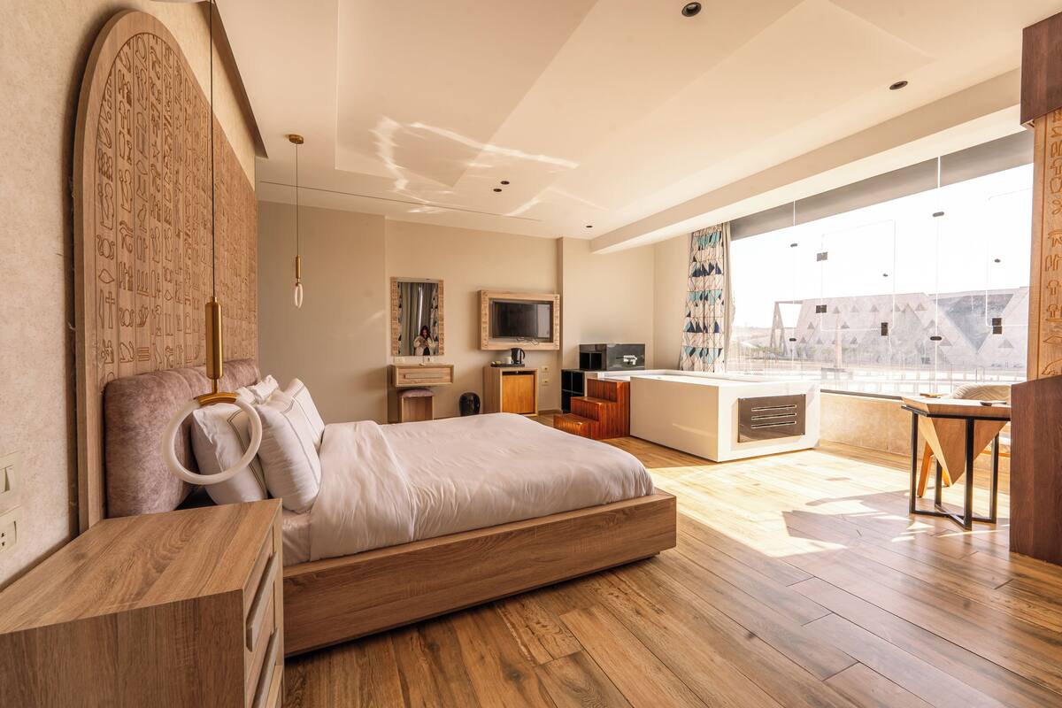 A modern bedroom with a large bed, a wooden headboard, a TV, and a balcony with a view.