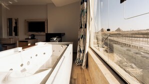 A modern bathroom with a large bathtub, a television, and a view of the pyramids. - Royal Great Pyramid INN (Giza)