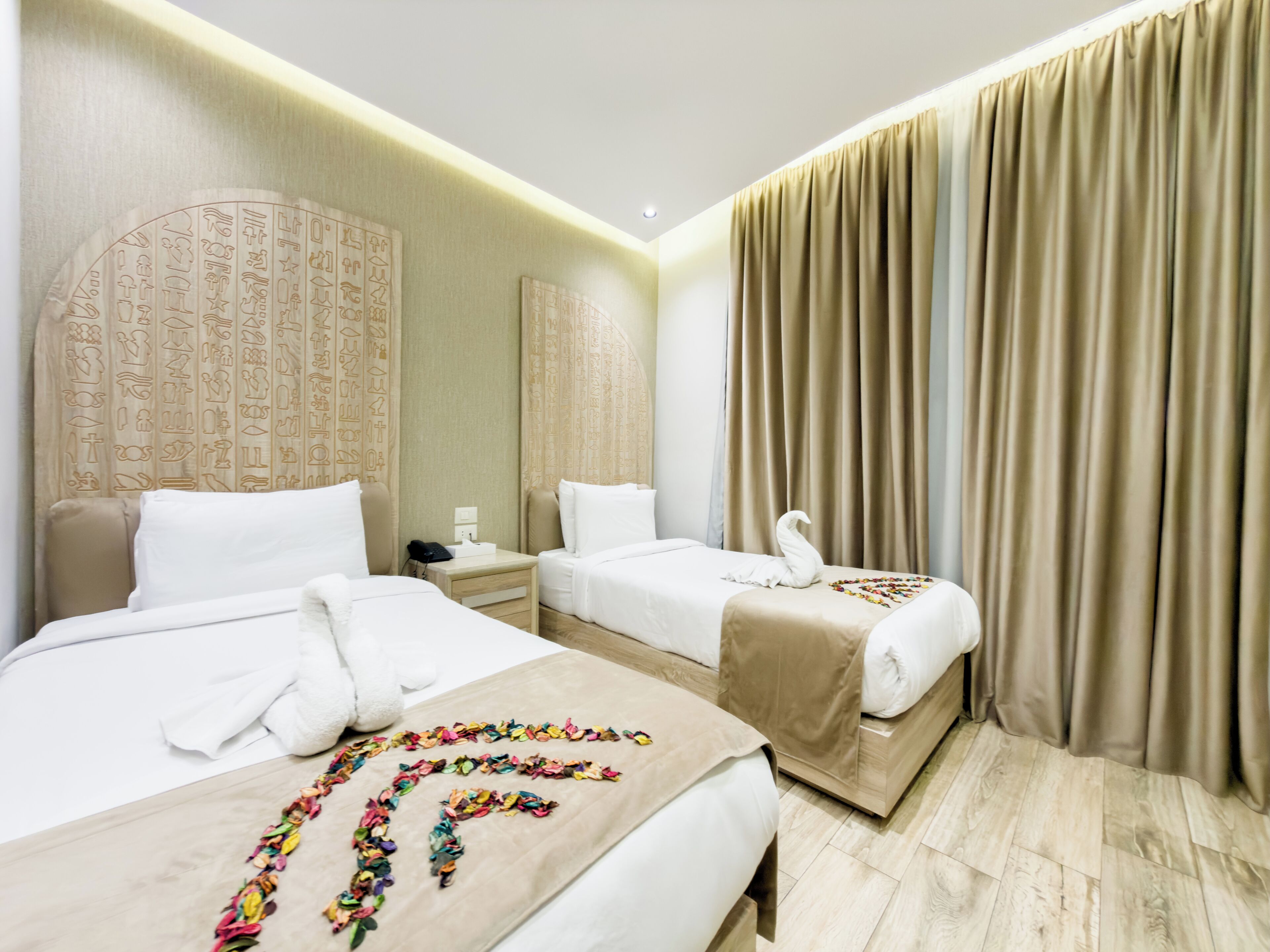 Ein Hotelzimmer mit zwei Betten, einem Nachttisch, einer Wand mit Hieroglyphen und einem großen Fenster mit Vorhängen.