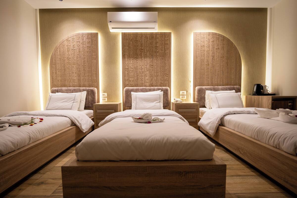 A hotel room with three beds, each with white linens and pillows, a wooden bench, and a wall with hieroglyphs.