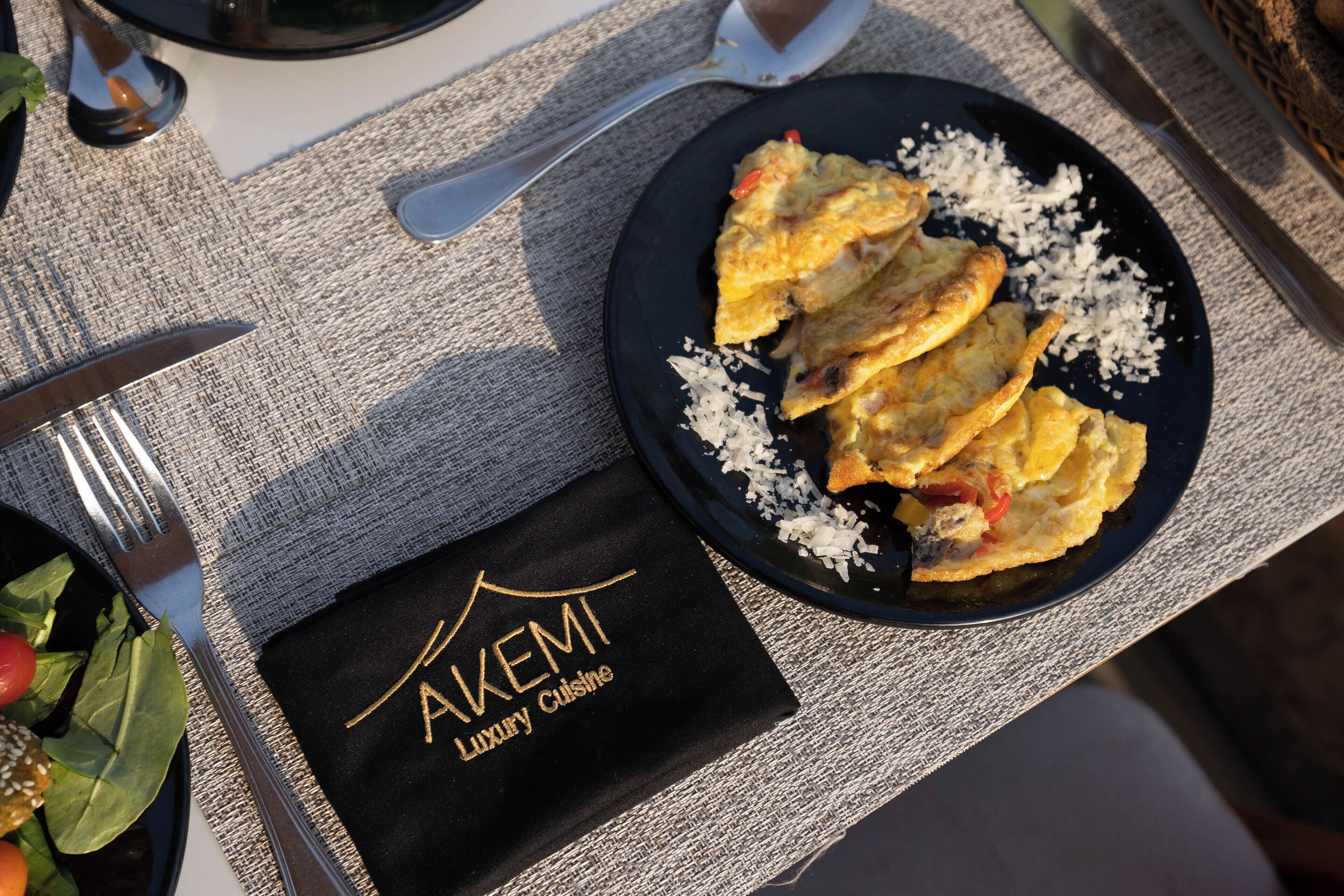 A table setting with a plate of food, a fork, a knife, and a napkin with "AKEMI Luxury Cuisine" text.