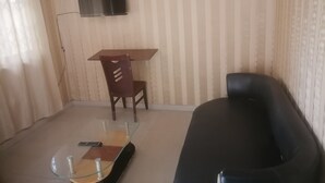 A living room with a black sofa, a glass coffee table, a wooden dining table with a chair, and a mounted television. - Vacation Inn Hotel (Accra)