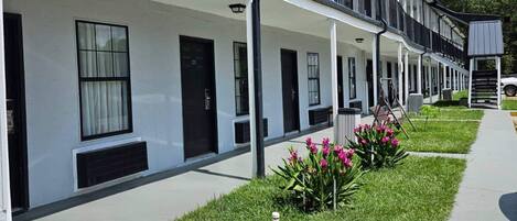 A row of modern, white buildings with black-framed windows and doors, a covered walkway, and a small garden with pink flowers.