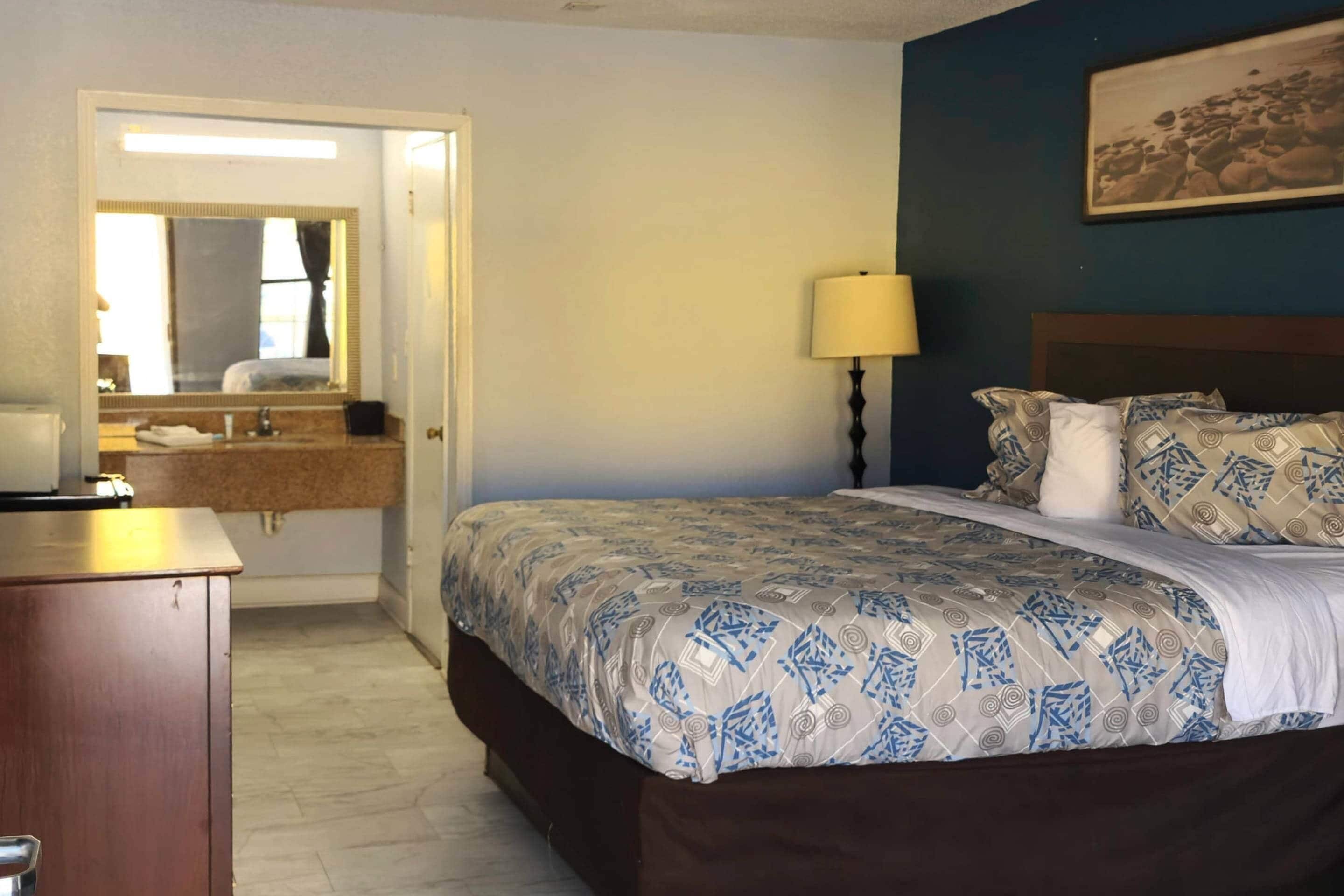 A bedroom with a bed, a nightstand, a lamp, a dresser, and a bathroom visible through an open door.