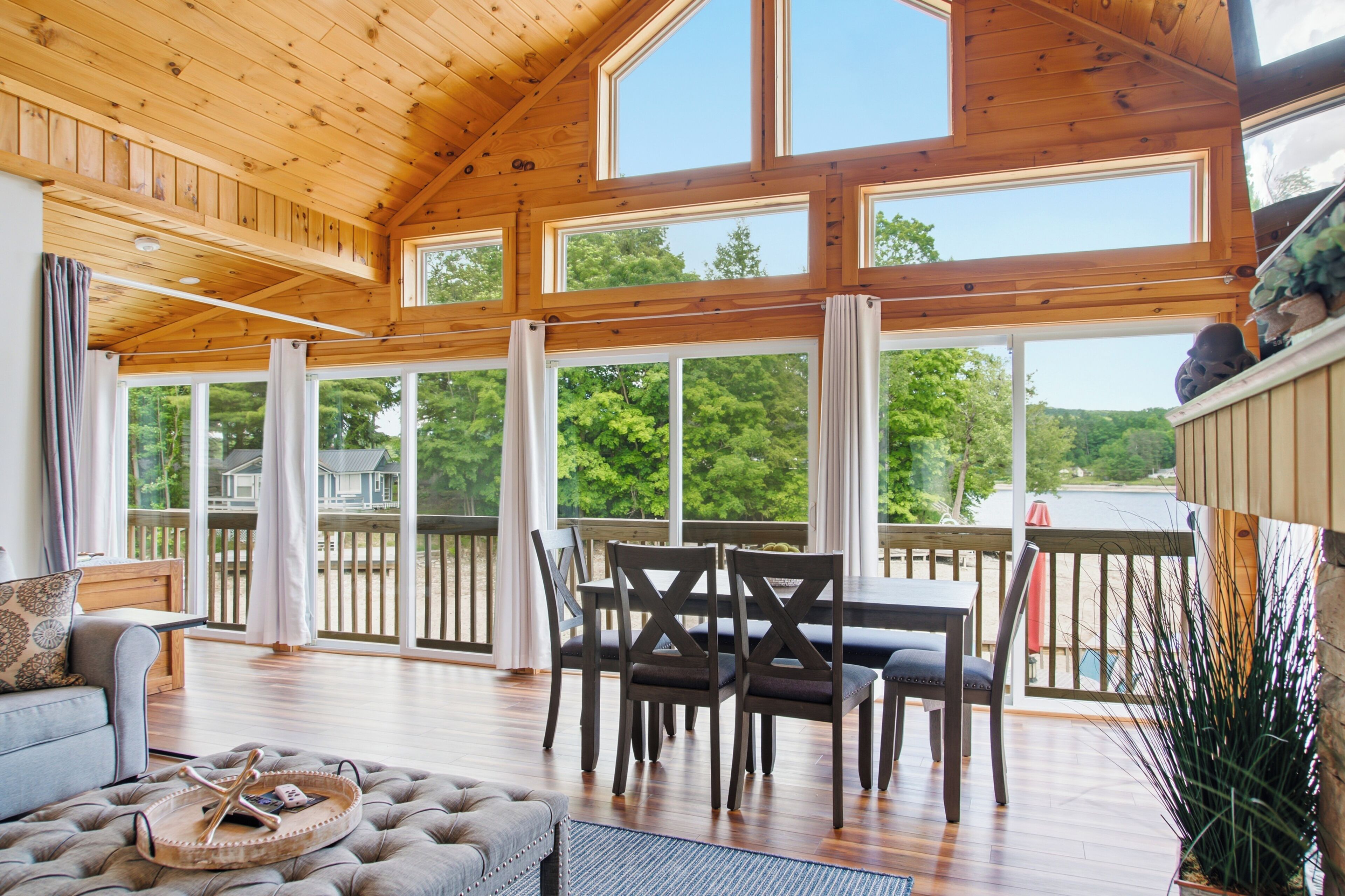 Amplia habitación con techo alto y ventanas grandes, terraza de madera y zona de comedor con sillas y mesa.