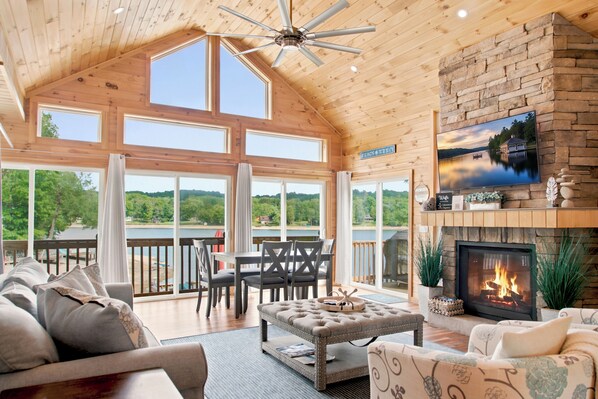 A spacious living room with a fireplace, a television mounted on a stone wall, and a large window overlooking a lake.