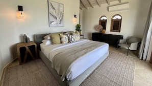 Ein Schlafzimmer mit Bett, Nachttisch, Wandleuchte, Deckenventilator, Kunstwerk und Blick ins Freie.