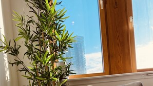 A modern room with a grey armchair, a wooden side table with a plant, and a view of a tall building through a window. - Spacious 2 Bedroom City pad in Old Street London (London)