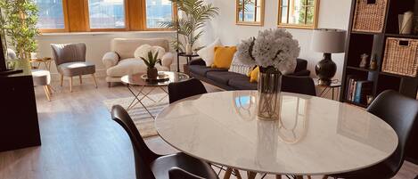 A modern living room with a round marble table, black chairs, a sofa with yellow cushions, and a bookshelf.