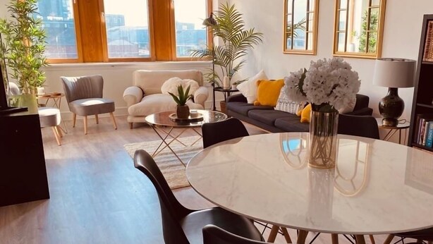 A modern living room with a round marble table, a sofa, and a chair. There are large windows with wooden frames, a potted plant, and a bookshelf. - Spacious 2 Bedroom City pad in Old Street London (London)