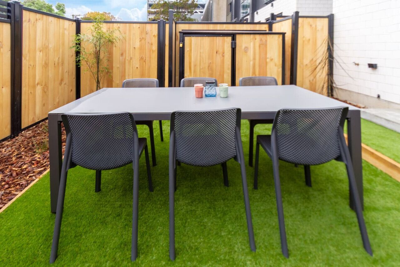 Une table rectangulaire entourée de quatre chaises, posée sur du gazon synthétique, le tout délimité par une clôture en bois.