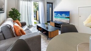 Una sala de estar moderna con un sofá, una mesa de centro y un televisor. Hay una planta y vistas al exterior a través de una ventana.