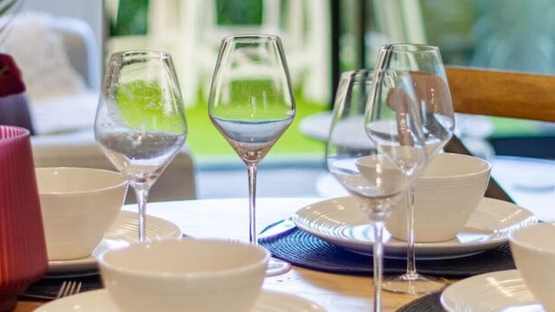 A dining table set with white plates, bowls, and wine glasses, with a view of a garden and outdoor seating area in the background.
