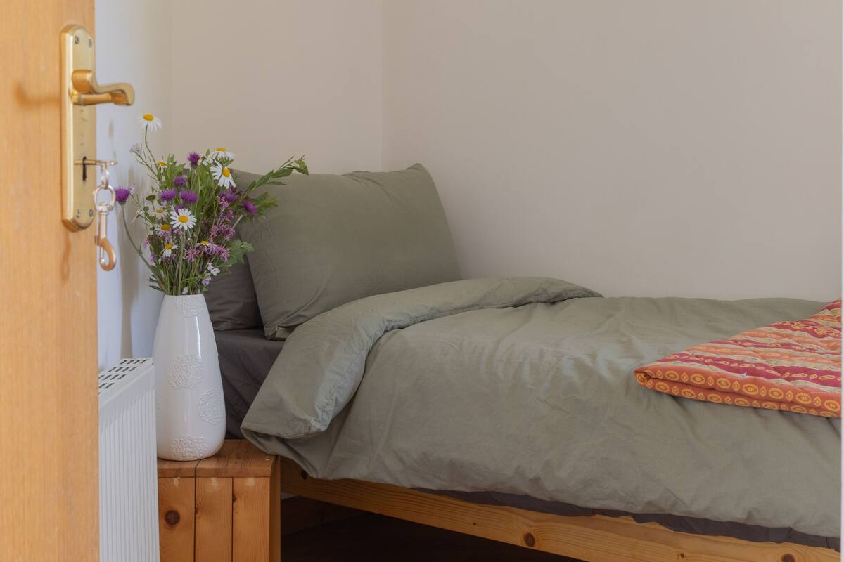 A single bed with a green blanket and a colorful throw, a white vase with flowers, and a wooden cabinet.