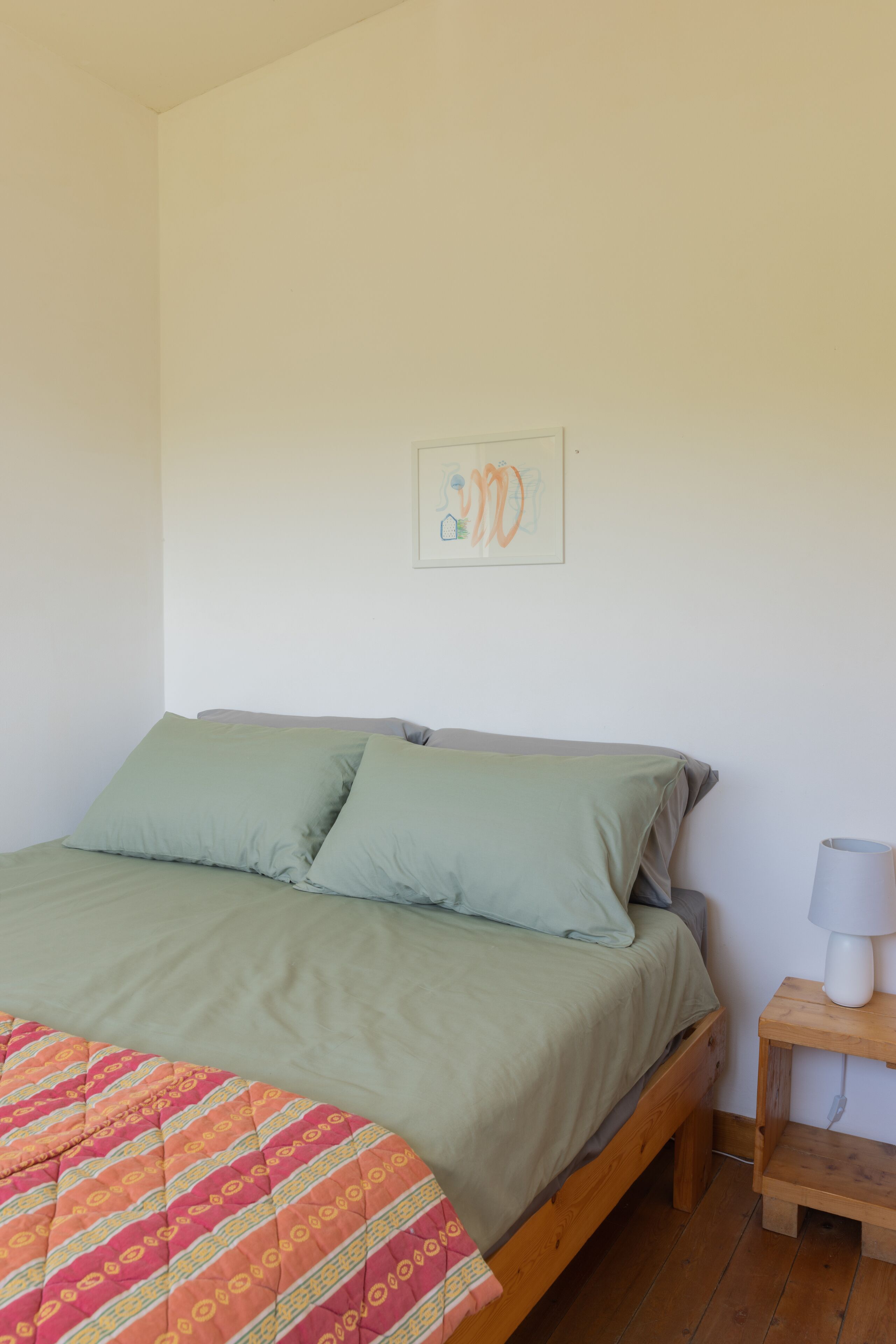 A single bed with a green bedspread and pillows, a wooden nightstand with a lamp, and a framed picture on the wall.