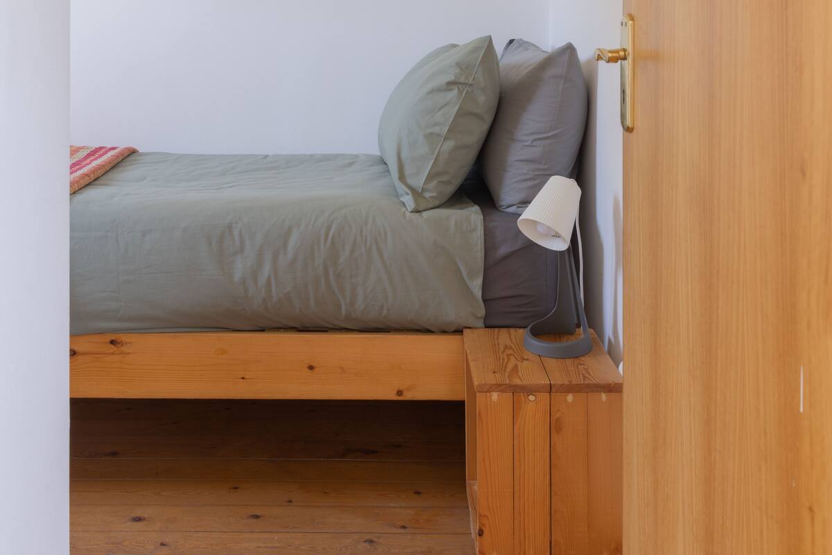 A single bed with a grey mattress and pillows, a bedside lamp, and a wooden nightstand.