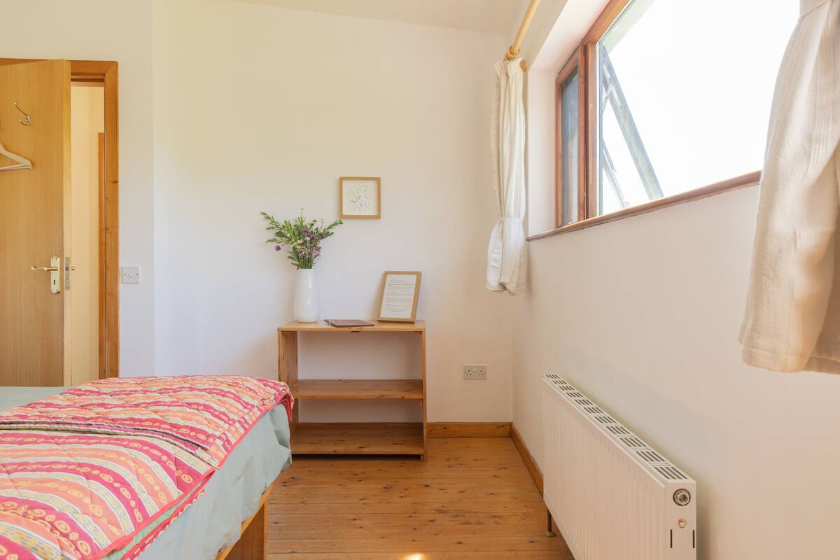 A small, cozy room with a bed, a wooden shelf with a vase and picture, a window with curtains, and a radiator.