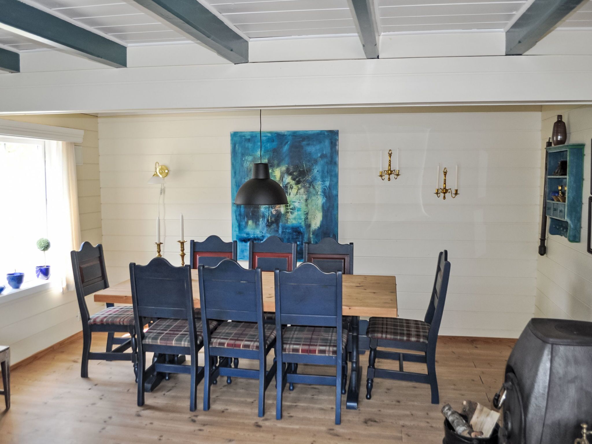 A dining area with a wooden table and chairs, a blue abstract painting on the wall, and a black pendant light.