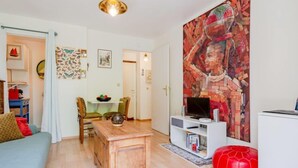 A living room with a wooden coffee table, a red ottoman, a TV on a white stand, and a colorful abstract painting on the wall. - Pavillon Gabriel by Interhome (Deauville-Trouville)