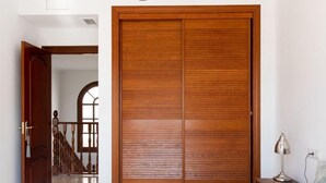 Une chambre avec une grande armoire en bois, un lit avec une literie blanche, une table de chevet avec une lampe, et une vue sur un escalier menant à une autre pièce.