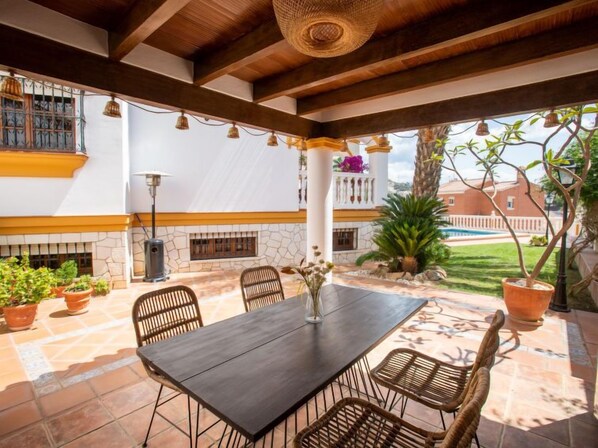 An outdoor patio with a wooden table and chairs, surrounded by potted plants and a view of a swimming pool.
