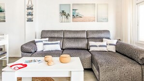 Een moderne woonkamer met een grijze modulaire bank, witte kussens, een witte salontafel met tijdschriften en gevlochten manden, en wanddecoratie met palmbomen en een strandscène.