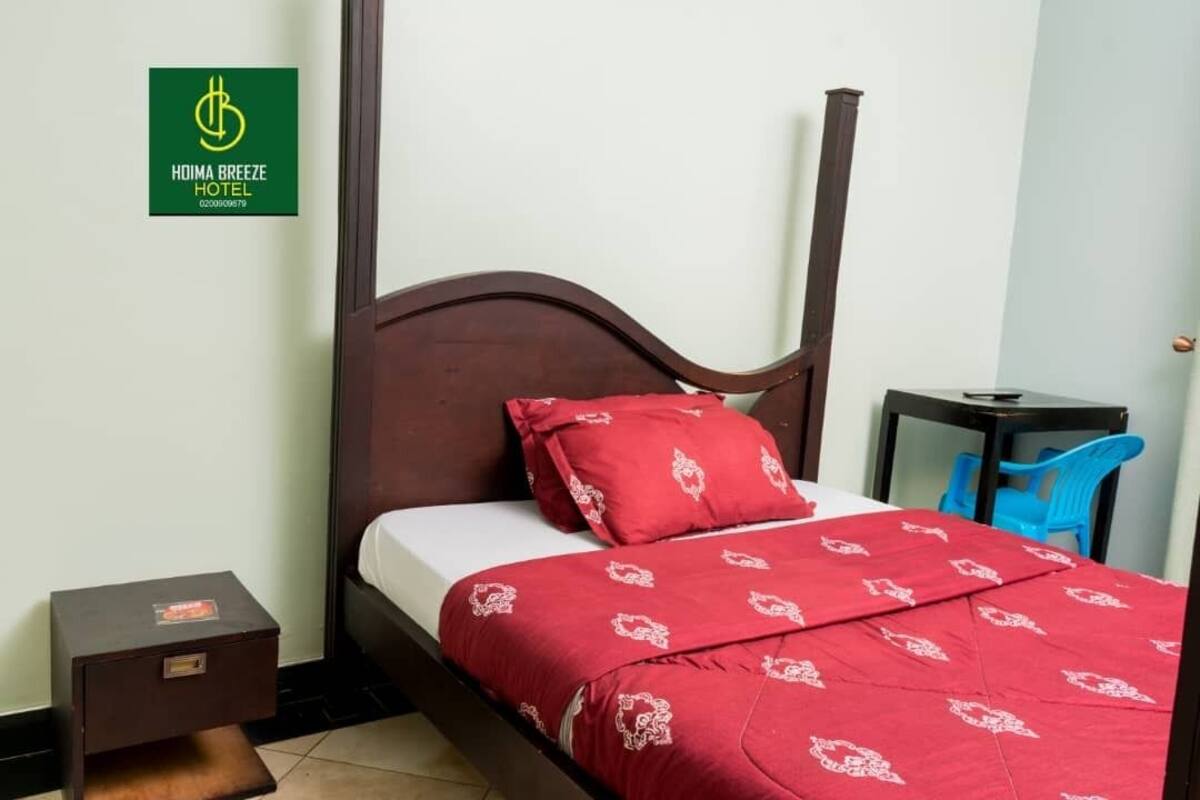 Une chambre d’hôtel avec un lit en bois, une literie rouge, une petite table de chevet, une chaise bleue et un panneau accroché au mur.