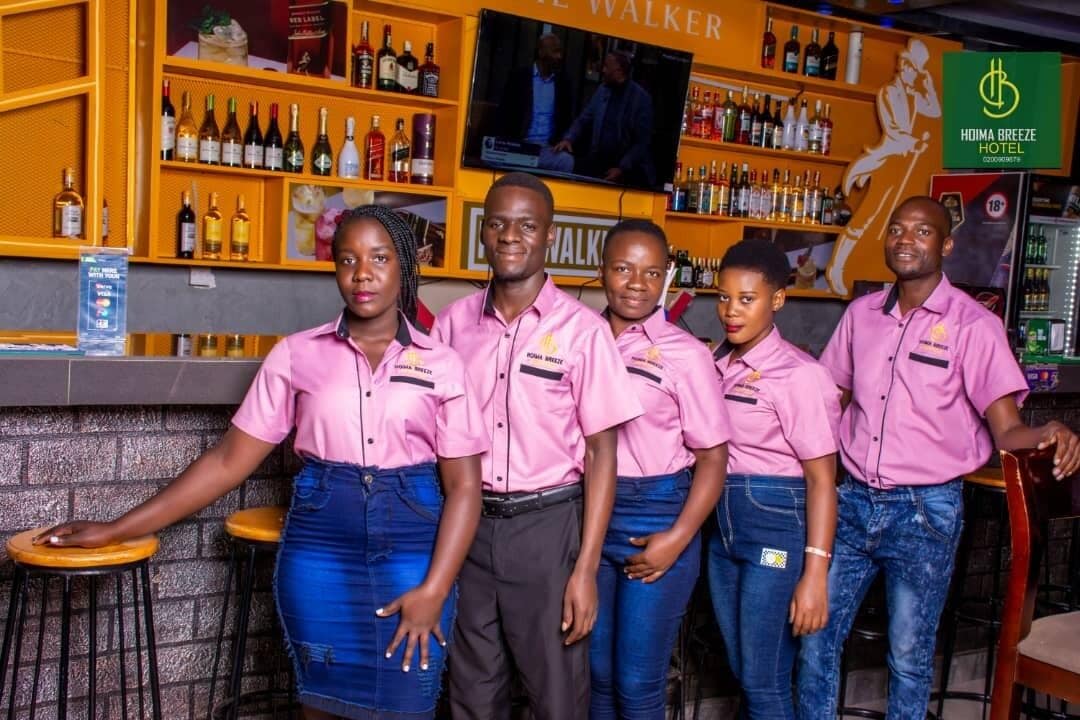 Un grupo de empleados de hotel con uniformes rosas de pie frente a una zona de bar con estantes llenos de botellas de licor y una pantalla de televisión.