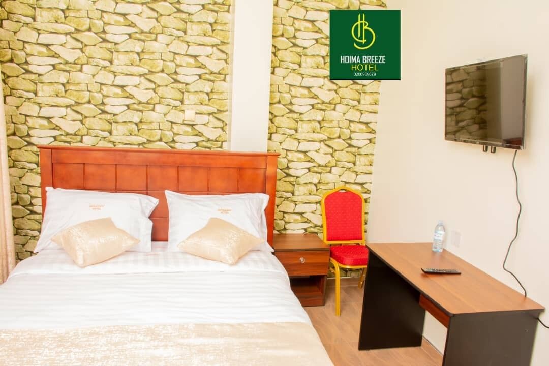 Ein Hotelzimmer mit Steinwand, einem Bett mit weißer Bettwäsche, einem roten Sessel, einem Schreibtisch und einem an der Wand befestigten Fernseher.