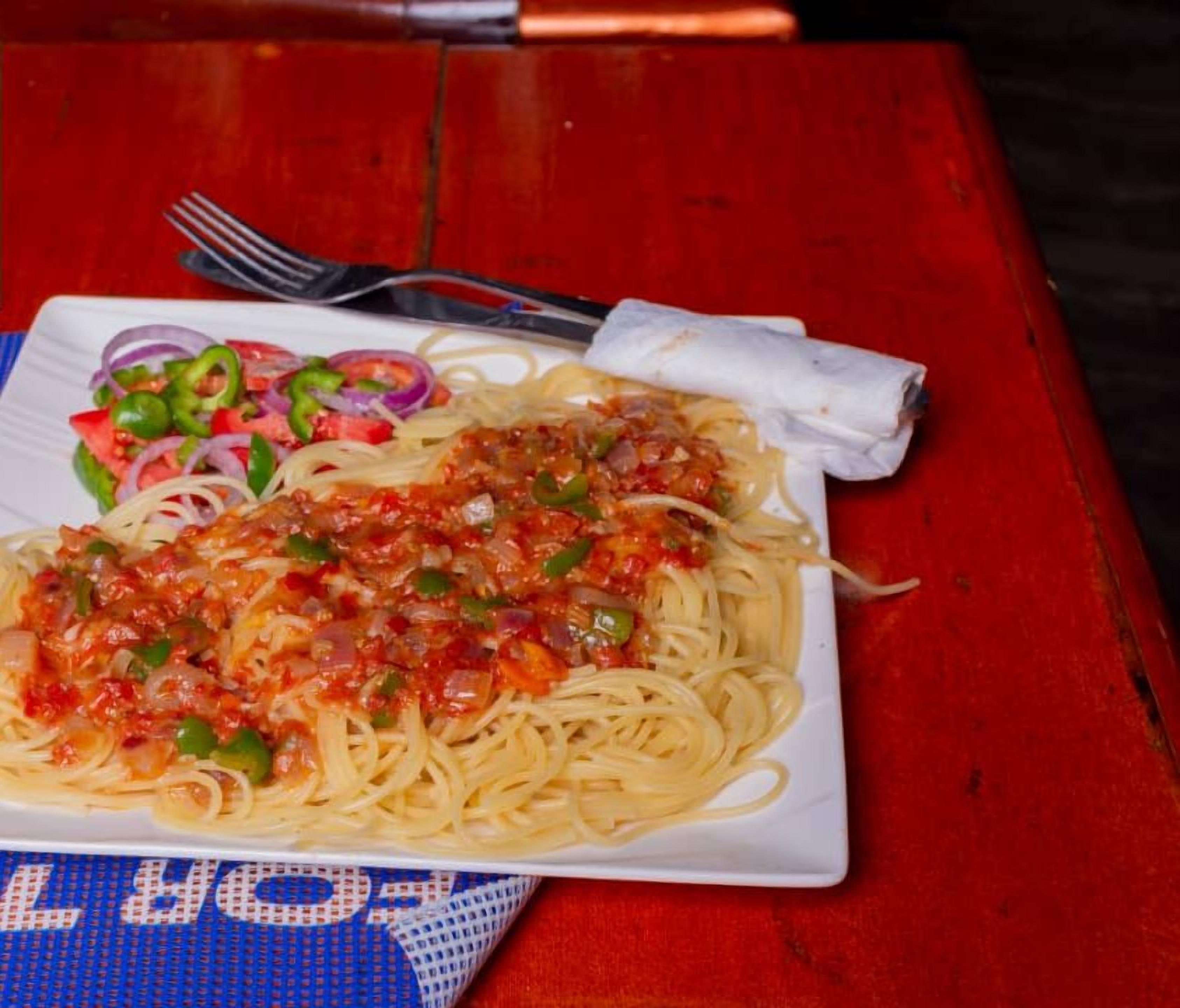 Een bord spaghetti met tomatensaus en groenten, een vork en een servet op een rode tafel met een blauwe tafelmat.