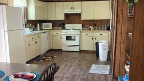 Een keuken met houten kastjes, een witte koelkast, een magnetron, een oven en een spoelbak. Er staat een eettafel met stoelen en een vloerkleed.