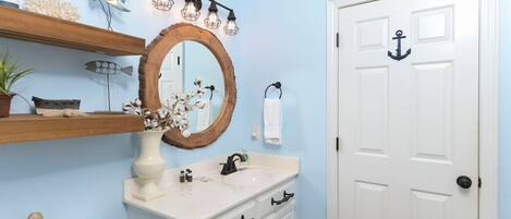 Un baño con una puerta blanca, un tocador de madera, un espejo grande y un estante con adornos.