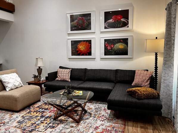 A living room with a dark grey sofa, a round coffee table, and a patterned rug. There are four framed floral pictures on the wall. - The Emerald at Mesa (Mesa)