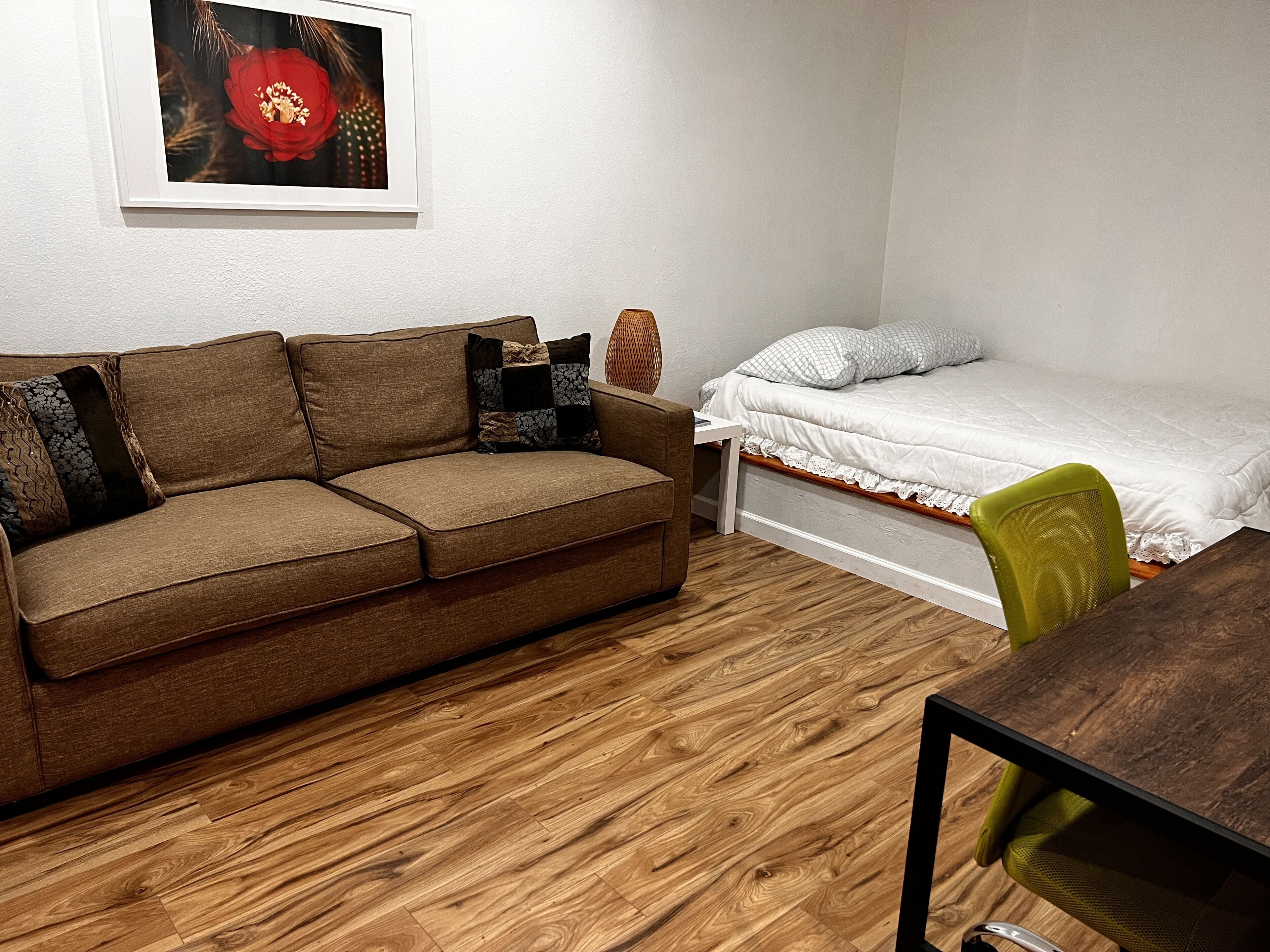 Una habitación pequeña y amoblada con un sofá marrón, una cama individual, piso de madera y una silla verde.