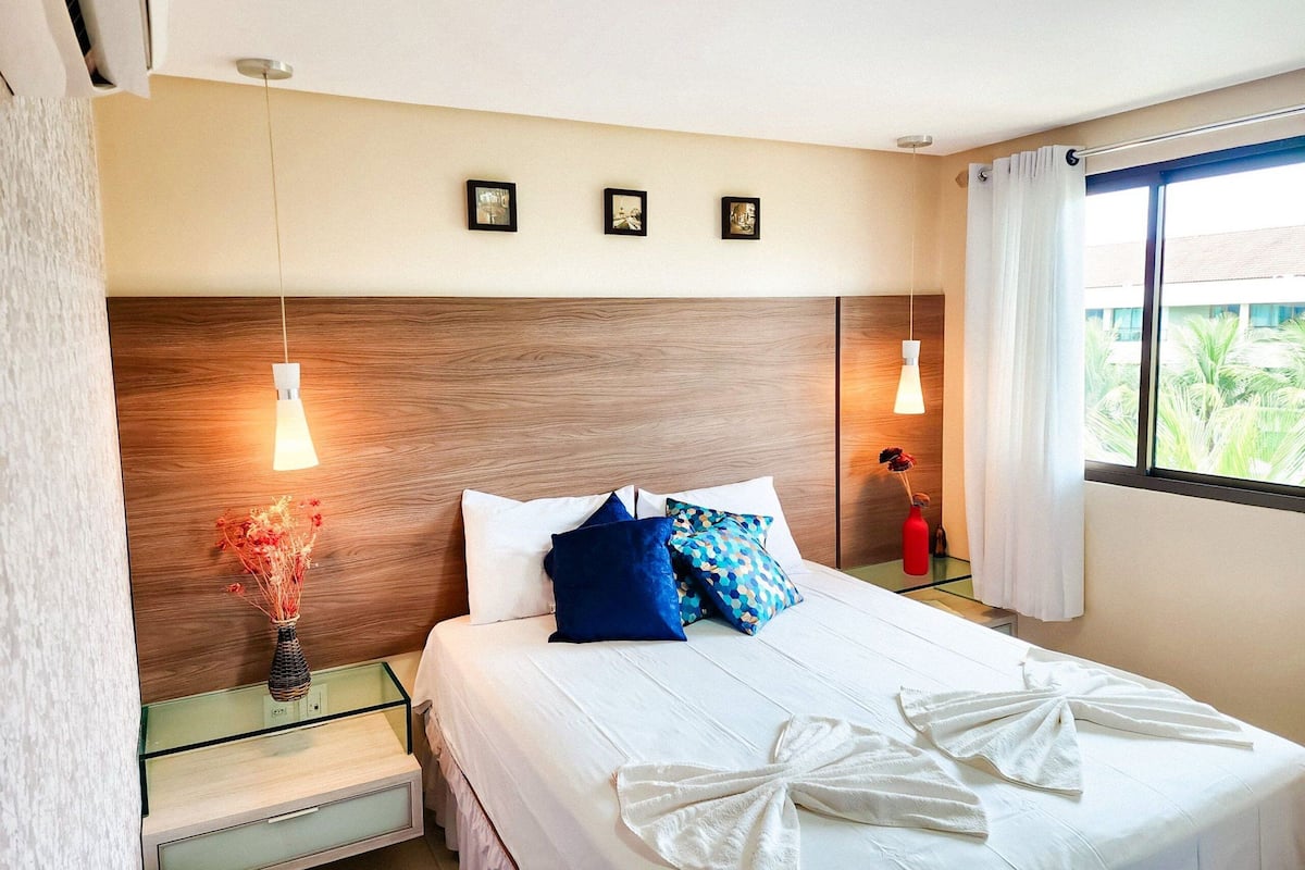 Ein Hotelzimmer mit Holz-Kopfteil, einem Bett mit weißer Bettwäsche und blauen Kissen, einem Nachttisch mit Blumenvase und einem Fenster mit Vorhängen.