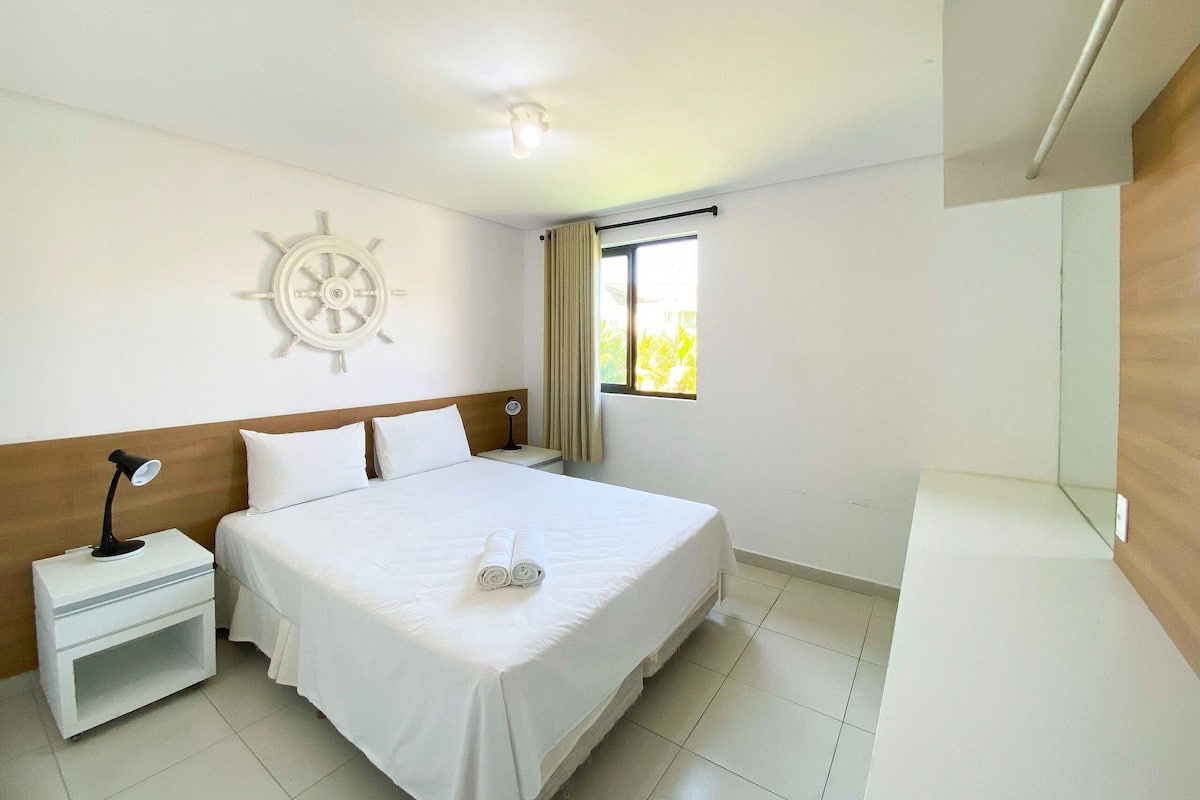 Ein Hotelzimmer mit Bett, Nachttisch, an der Wand montiertem Steuerrad, Fenster mit Vorhängen und Deckenleuchte.