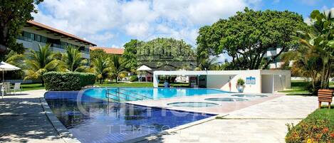 A swimming pool with a tiled deck, surrounded by palm trees and a building with a red roof.