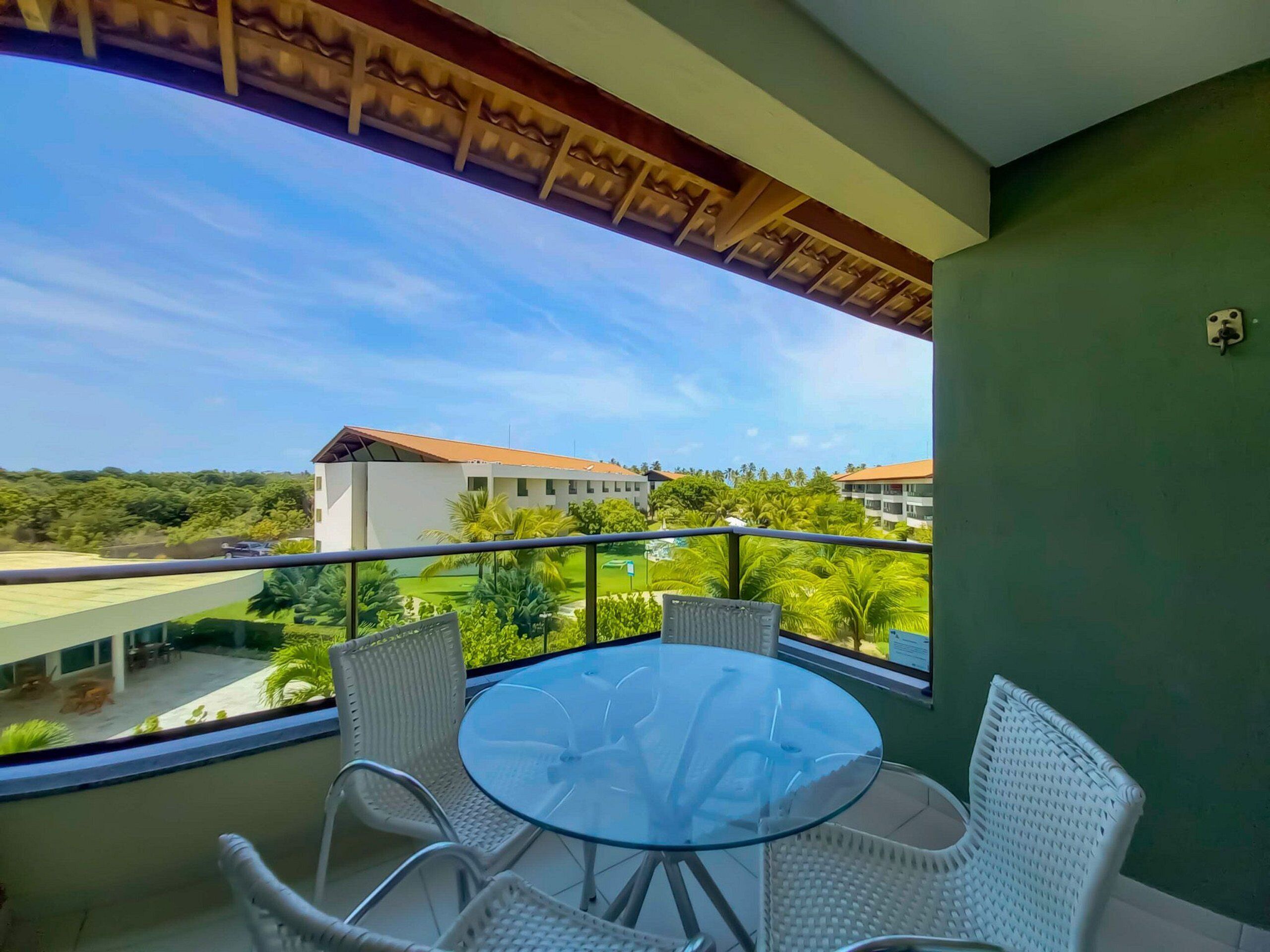 A balcony with a glass table and white wicker chairs, overlooking a building with a red roof and greenery.