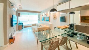 A modern kitchen with a glass dining table, a sofa, and a TV mounted on the wall. - Flats à beira-mar no Carneiros Beach Resort (Tamandaré)