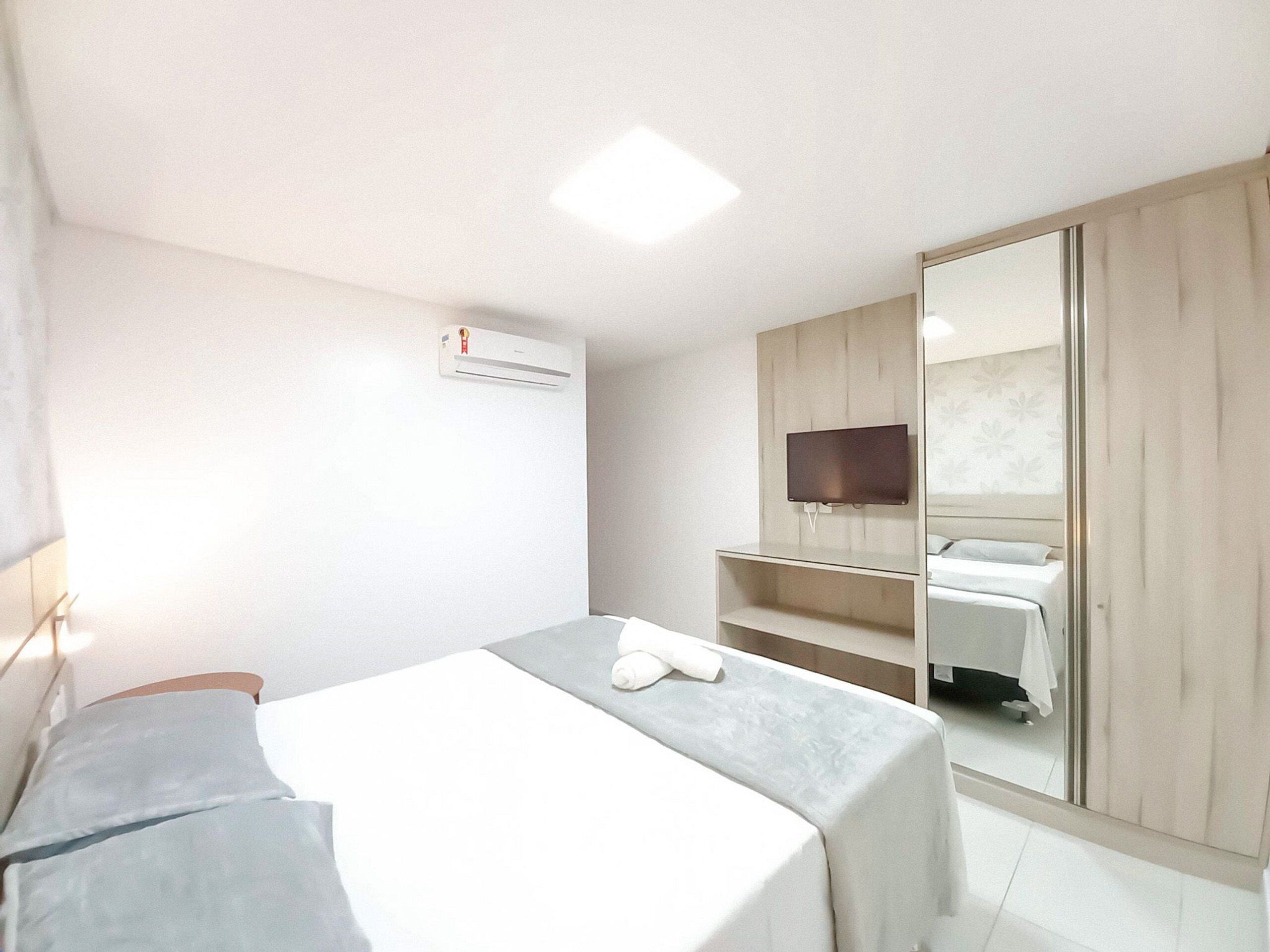 Quarto de hotel com cama, televisão fixada em um painel de madeira, espelho e ar condicionado.