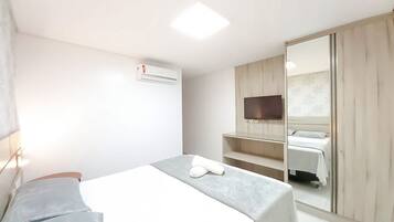 Quarto de hotel com cama, televisão fixada em um painel de madeira, espelho e ar condicionado.
