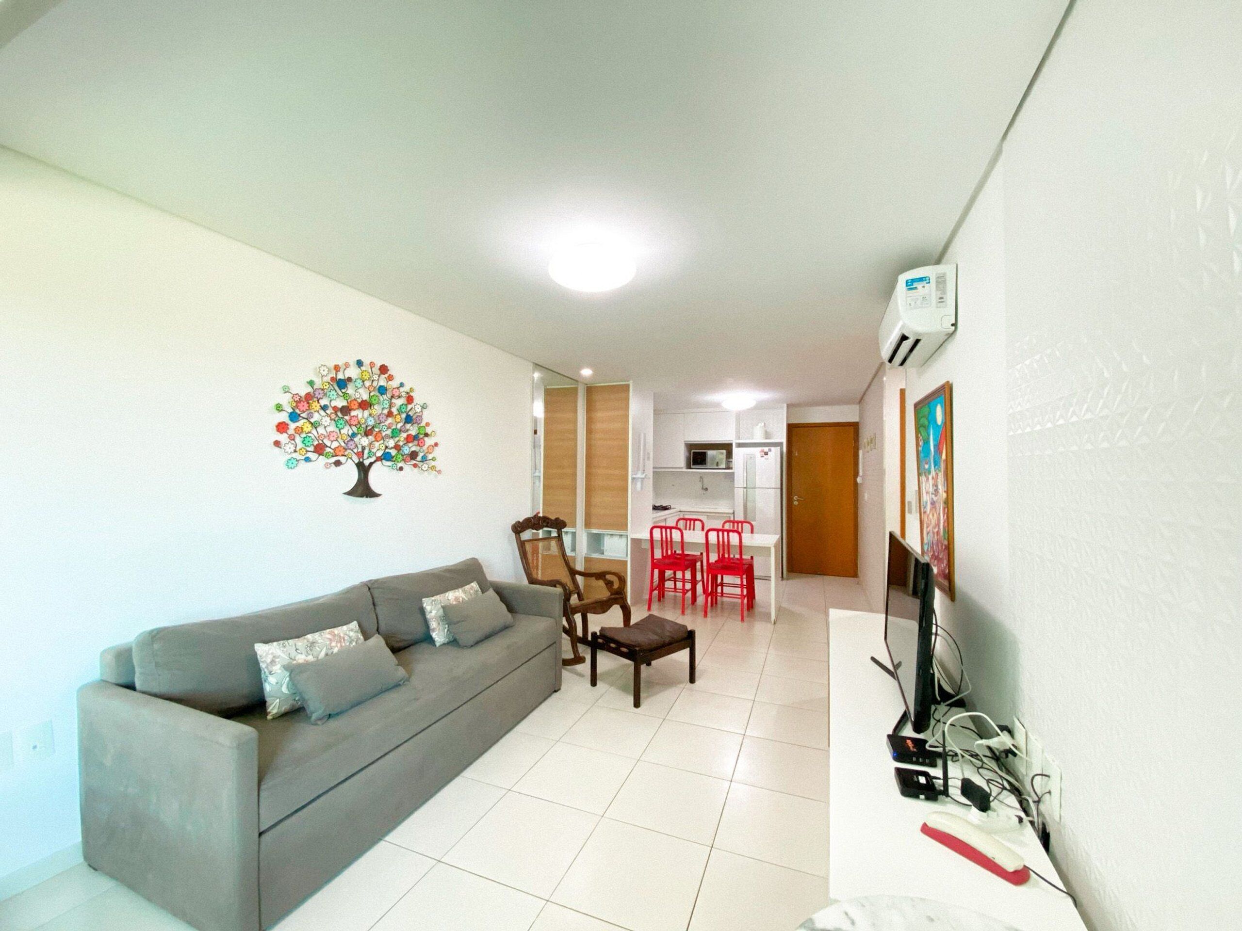 A living room with a sofa, a coffee table, and a dining area with red chairs. There is a kitchen in the background with white cabinets and a microwave.