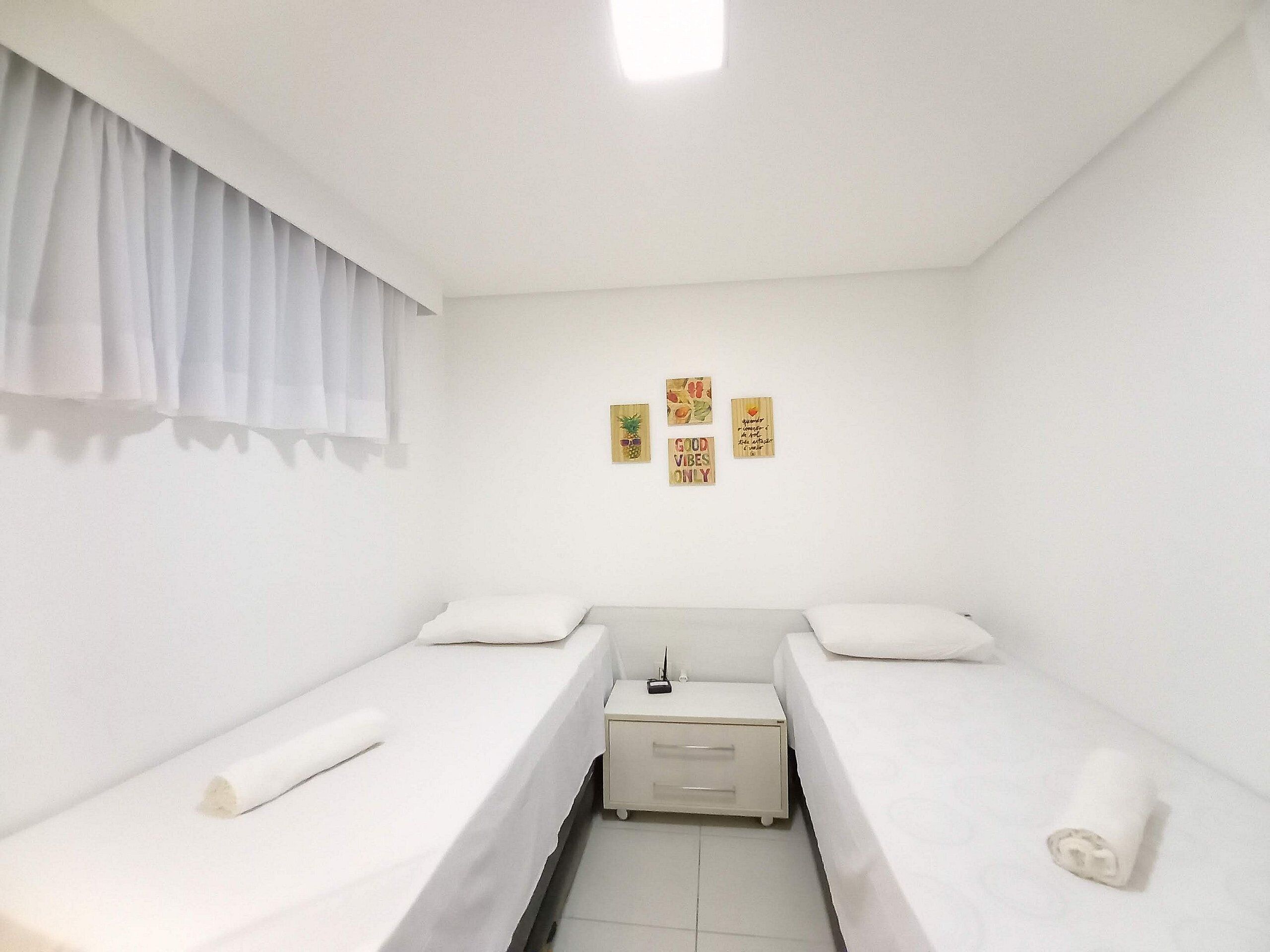 A small, clean room with two single beds, a bedside table, and a wall-mounted shelf with decorative items.